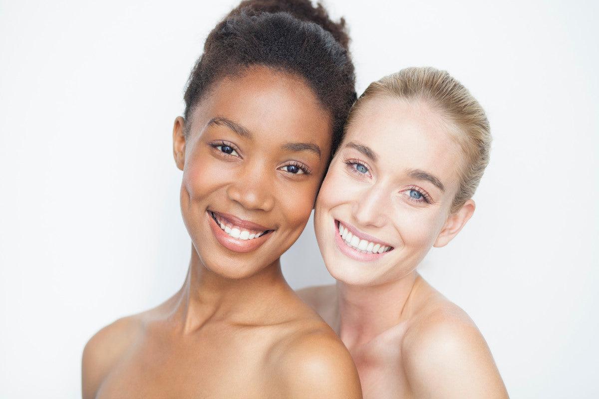 Two smiling women with glowing skin and bare shoulders, standing close together and looking at the camera against a white background.