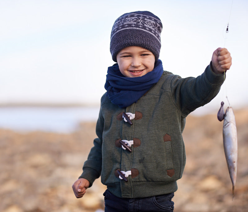 Smiling young boy wearing a knit hat and scarf, proudly holding up a small fish he caught on a rocky shoreline.
