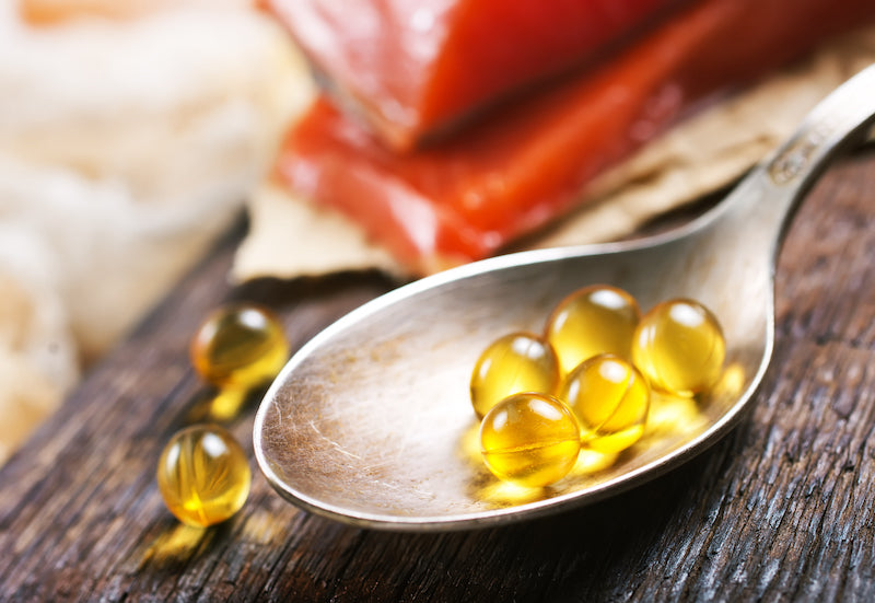 Close-up of omega-3 fish oil capsules on a spoon, with raw salmon slices in the background on a rustic wooden table.