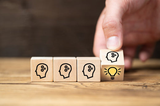 Hand arranging wooden blocks with head icons; the final block features a glowing lightbulb inside the head, symbolizing innovation or a bright idea.