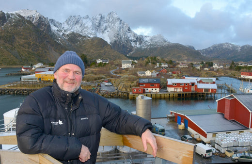 Smiling man in a winter jacket and beanie standing on a wooden deck with a view of a Nordic fishing village, red houses, fjord, and snow-covered mountains in the background.
