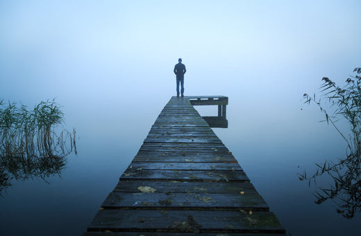 Silhouette of a person standing alone at the end of a wooden pier surrounded by still water and dense fog, creating a serene and introspective atmosphere.