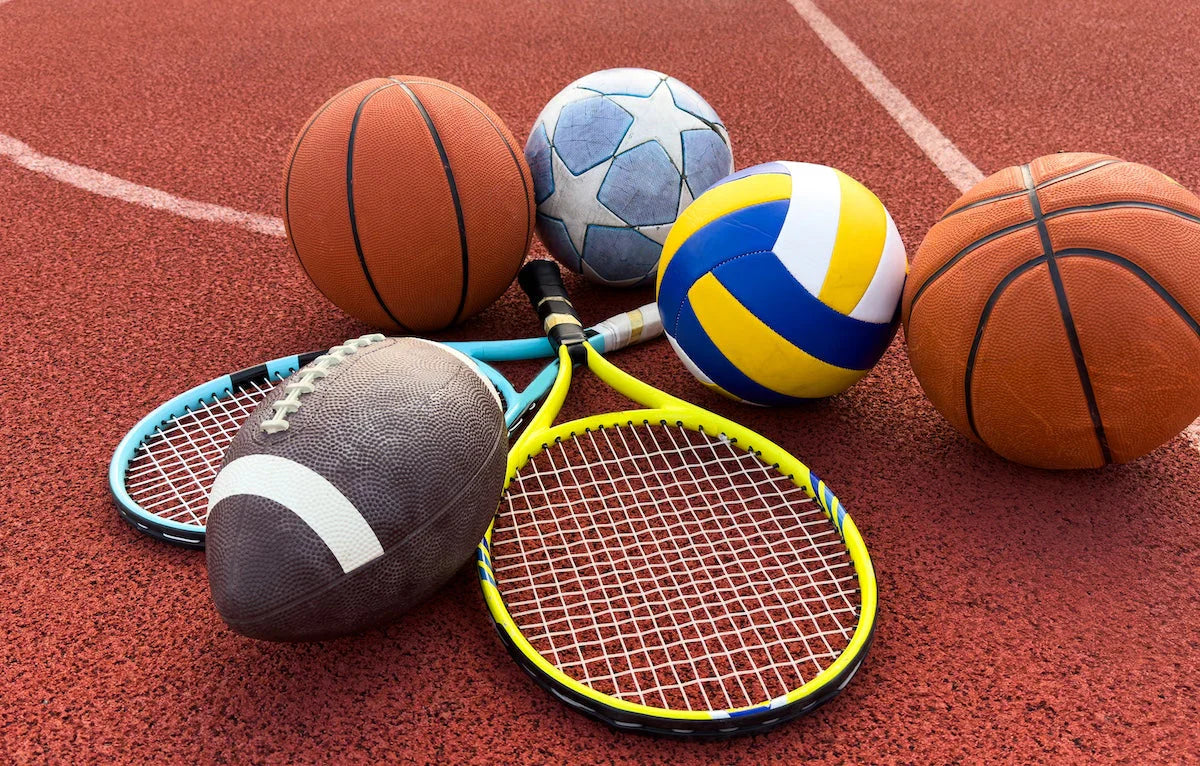Collection of sports equipment including basketballs, a soccer ball, a volleyball, an American football, and tennis rackets on a red court surface.