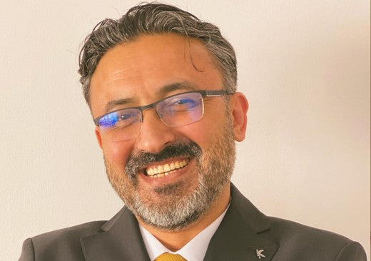 Smiling man with salt-and-pepper hair and beard, wearing glasses, a dark suit, white shirt, and a yellow tie, standing in front of a light-colored wall.