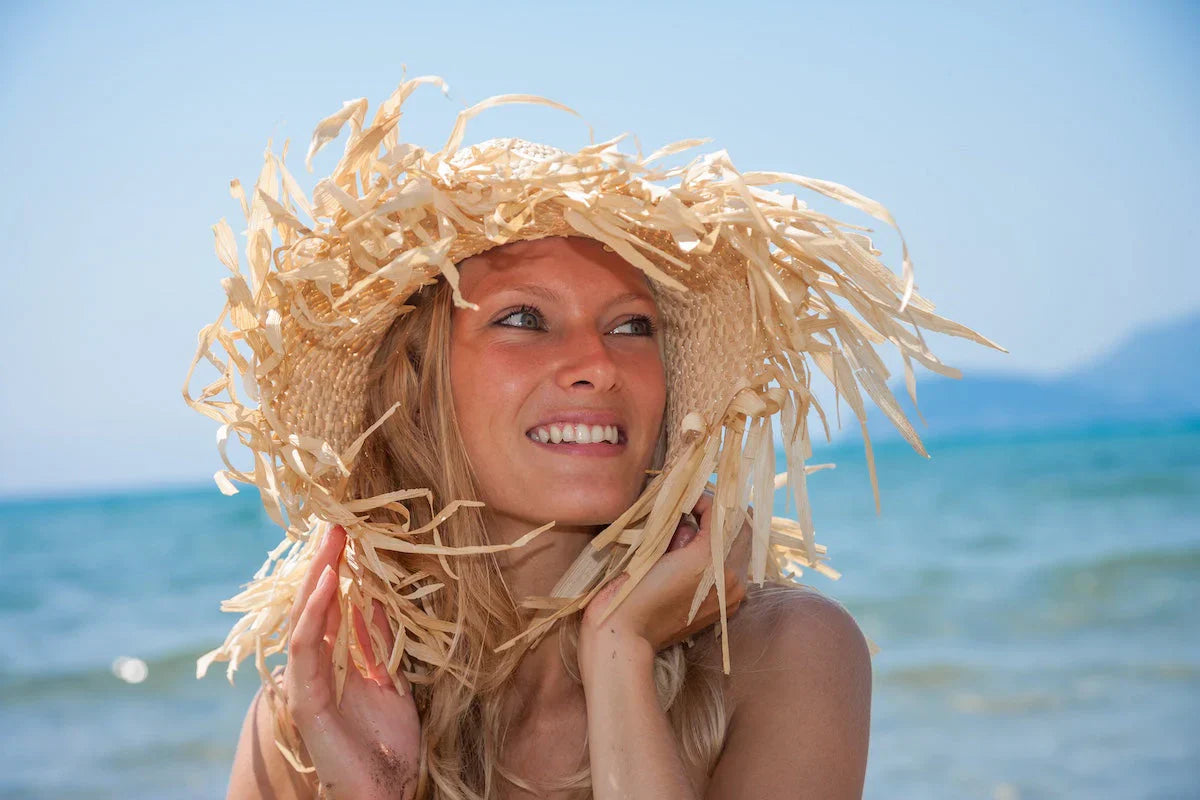 A smiling woman wearing a large straw hat, standing on a beach with the ocean and mountains in the background on a sunny day.