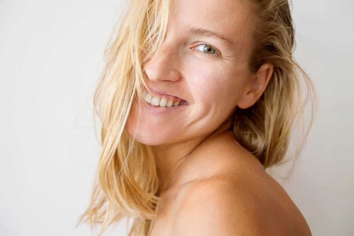 Close-up of a smiling blonde woman with glowing skin and natural makeup, looking over her shoulder with hair partially covering her face.