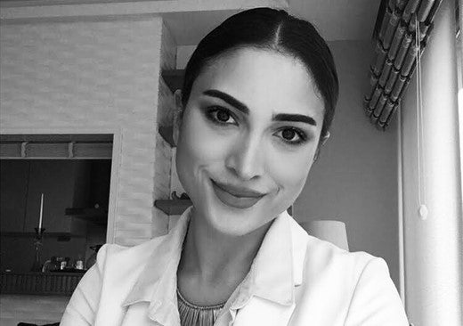 Black and white portrait of a woman with dark hair tied back, wearing a white coat and smiling confidently in a well-lit room with modern decor.