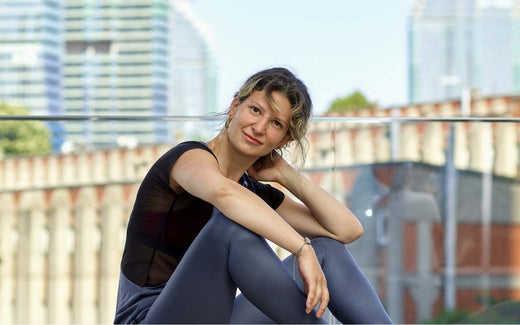 Young woman in athletic wear sitting outdoors on a balcony or terrace with city skyscrapers and a brick viaduct in the background, smiling thoughtfully with one hand resting on her neck.