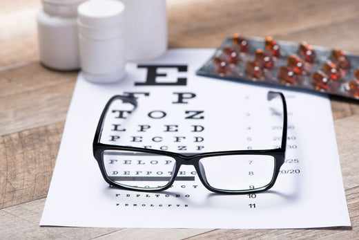 Eyeglasses placed on a vision test chart with medicine bottles and blister packs of capsules in the background, representing eye health and vision care.