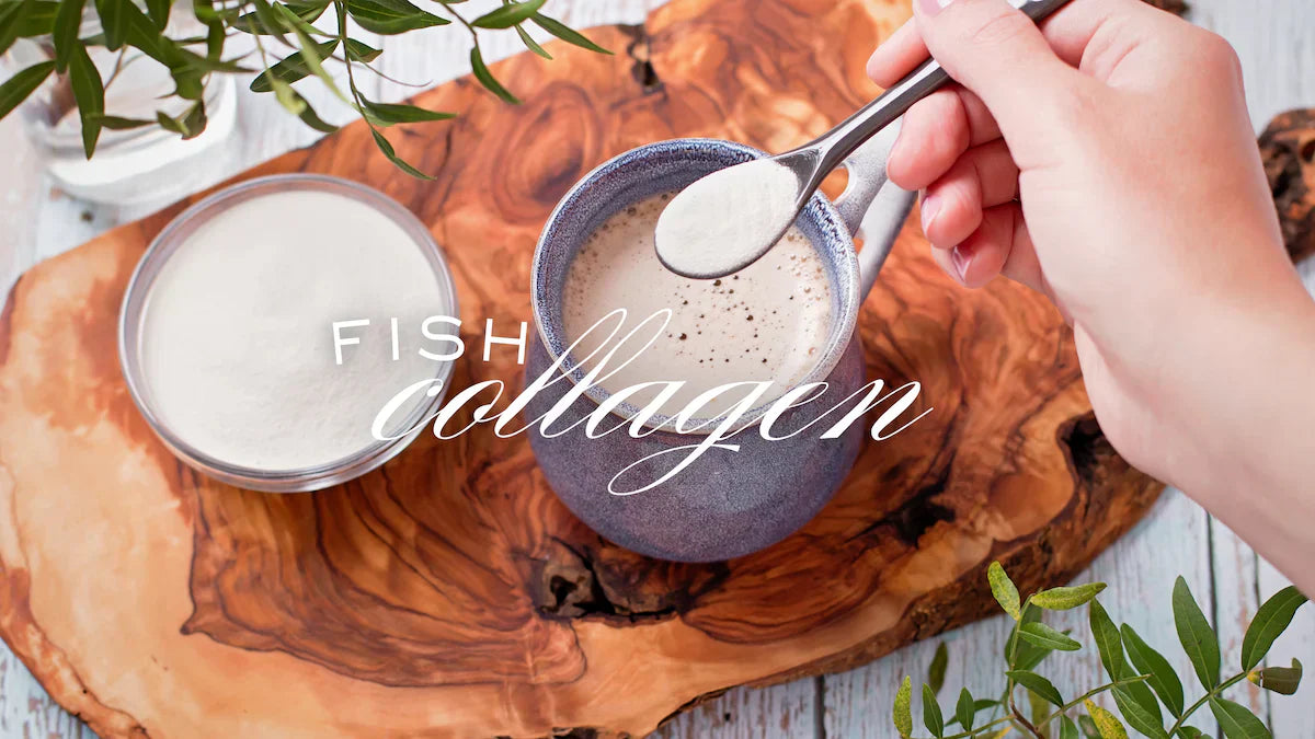 A hand adding a spoonful of fish collagen powder into a mug of coffee, with a bowl of white collagen powder on a wooden board in the background.