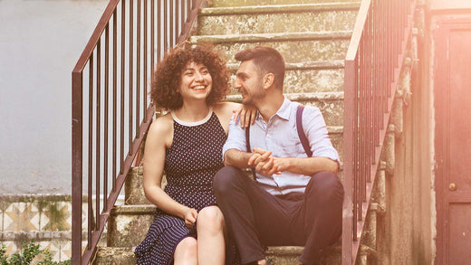 Smiling couple sitting on outdoor stairs, looking at each other affectionately, with warm sunlight illuminating the scene.