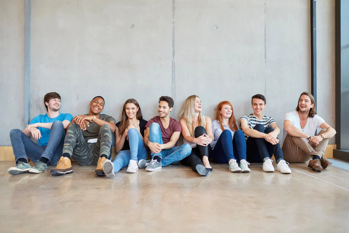 Group of diverse young students sitting on the floor against a wall, smiling and talking together.