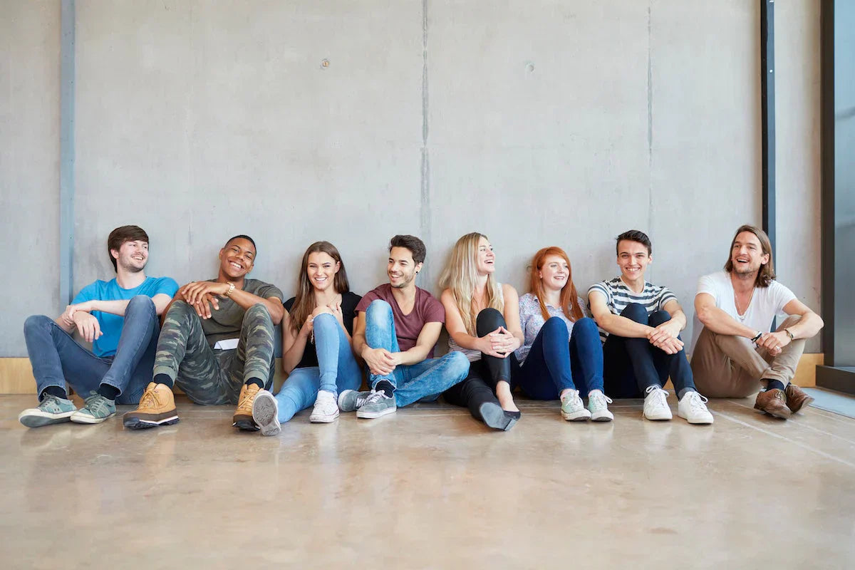 Group of diverse young students sitting on the floor against a wall, smiling and talking together.