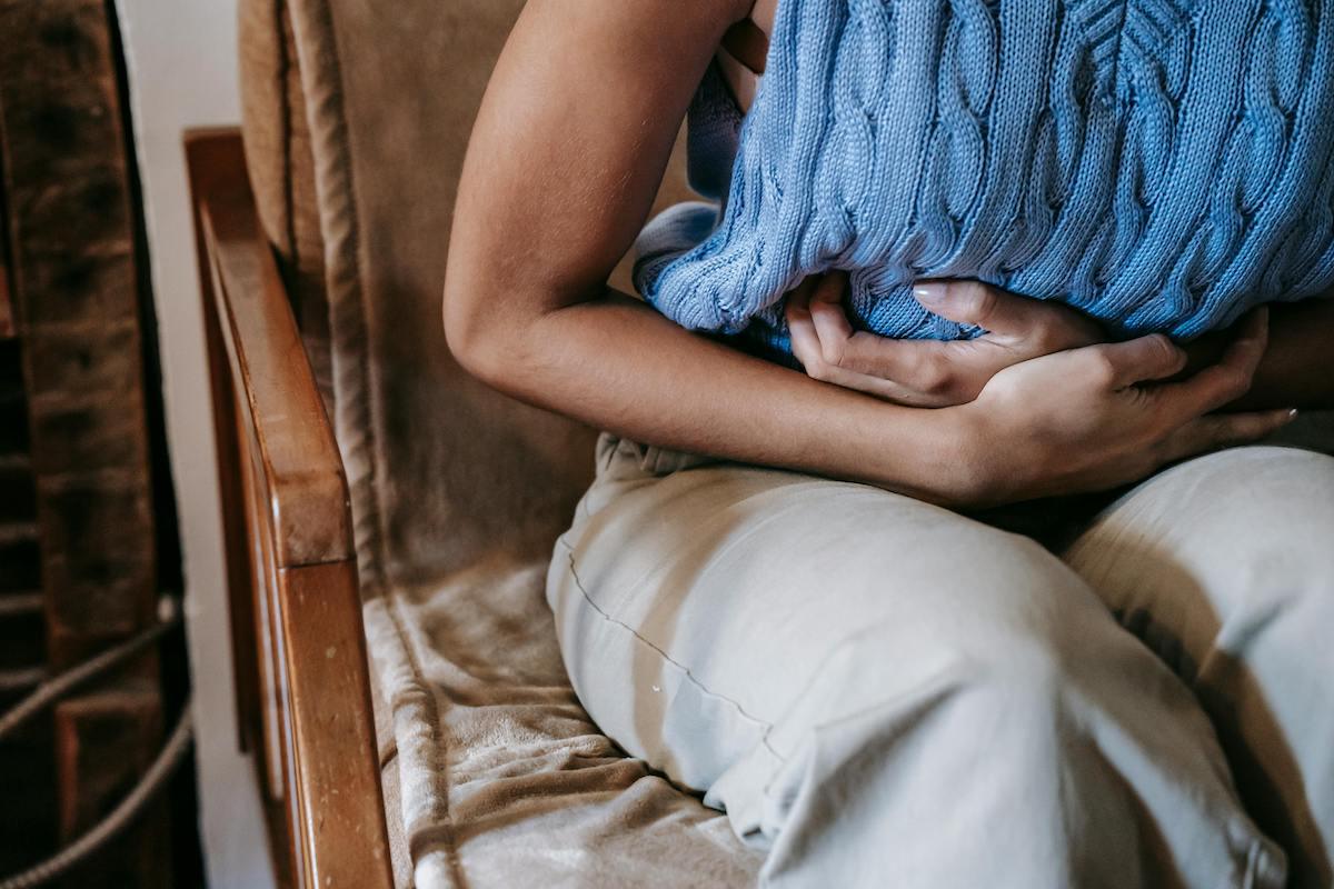 Person sitting on a couch holding a blue knitted pillow against their stomach, appearing to experience abdominal discomfort or pain.
