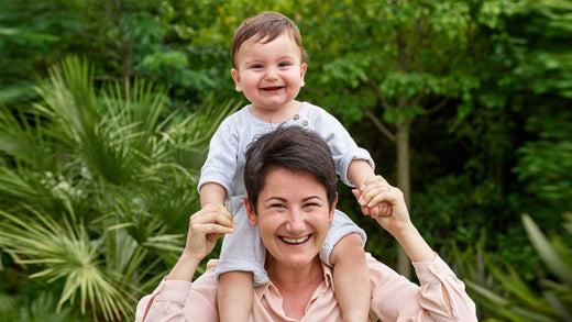 Smiling woman outdoors holding a happy baby on her shoulders, surrounded by lush green foliage and tropical plants.