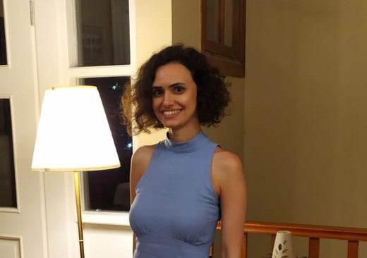 Smiling woman with curly dark hair wearing a sleeveless light blue dress, standing indoors near a lit floor lamp and a window at night.