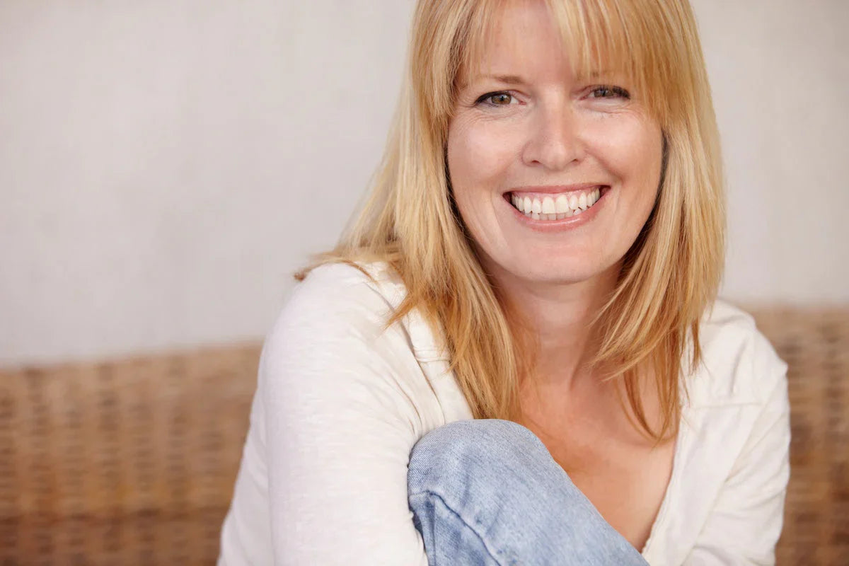 Smiling middle-aged woman with blonde hair sitting casually in a white top and jeans.