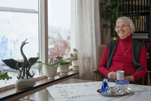 Elderly woman with glasses sitting in a cozy living room by the window, smiling while holding a cup of tea, surrounded by houseplants and decorative items.