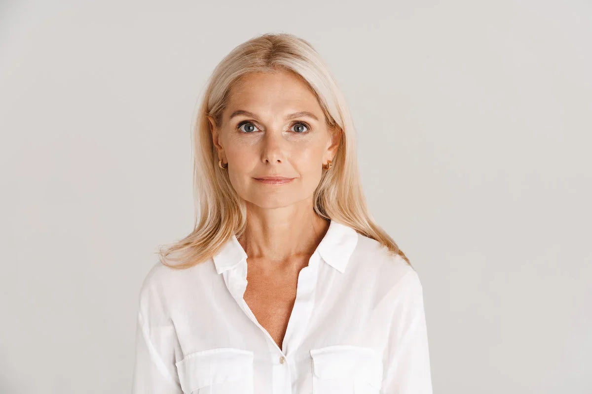 A mature blonde woman with long hair wearing a white shirt, standing against a neutral background and looking confidently at the camera.