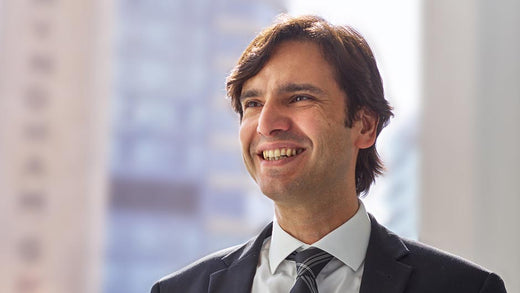 Confident smiling businessman in a suit and tie, standing in front of a blurred office window with cityscape in the background.