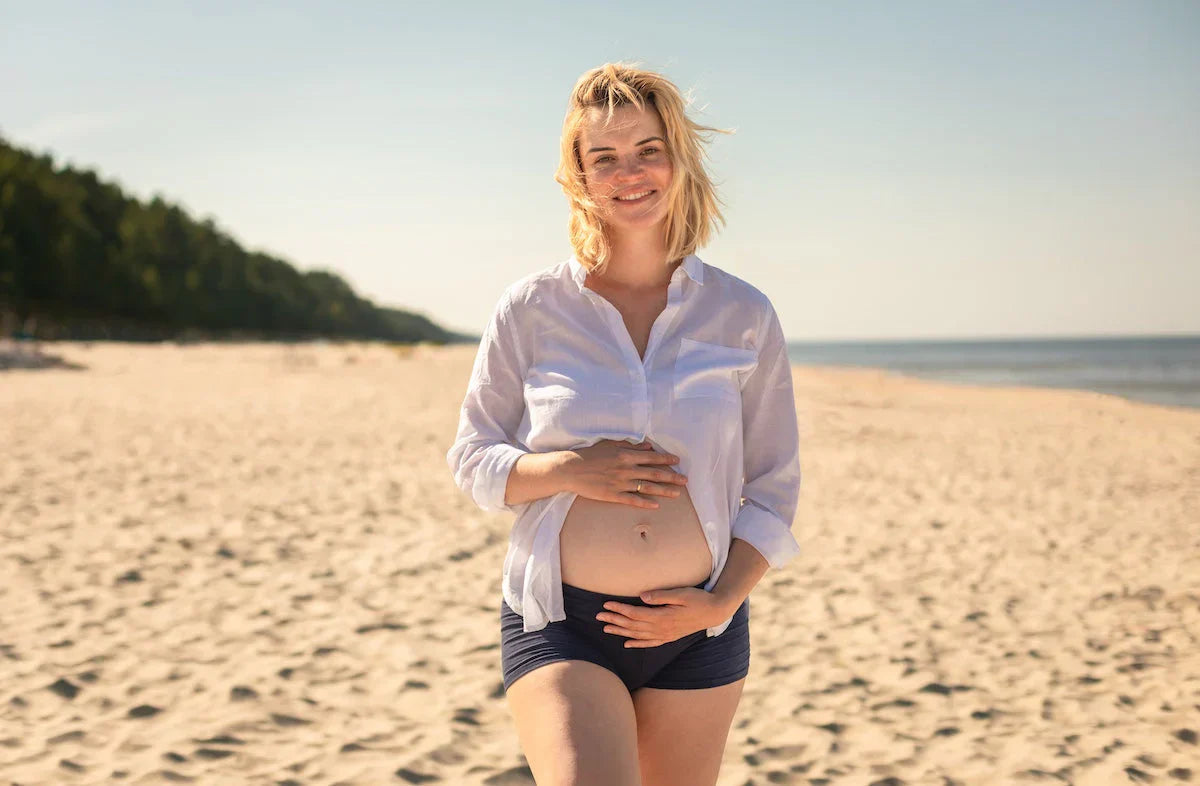 Smiling pregnant woman in a white shirt and shorts touching her belly while walking on a sandy beach.