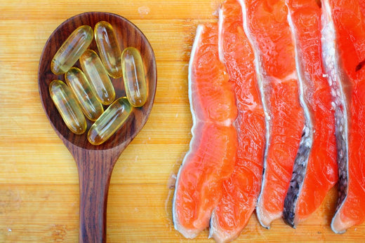 Wooden spoon with omega-3 fish oil capsules next to raw salmon fillets on a wooden cutting board, symbolizing natural sources of healthy fats.