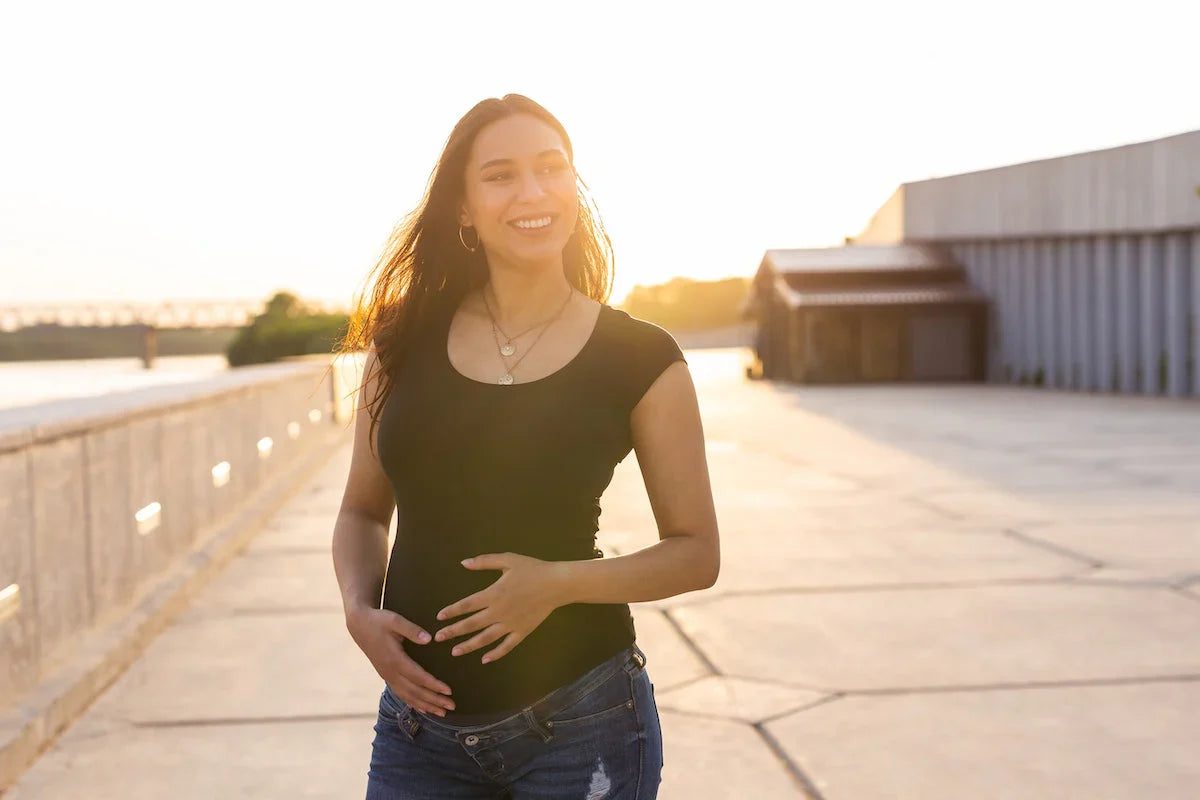 Smiling pregnant woman in casual clothes holding her belly while walking outdoors at sunset.