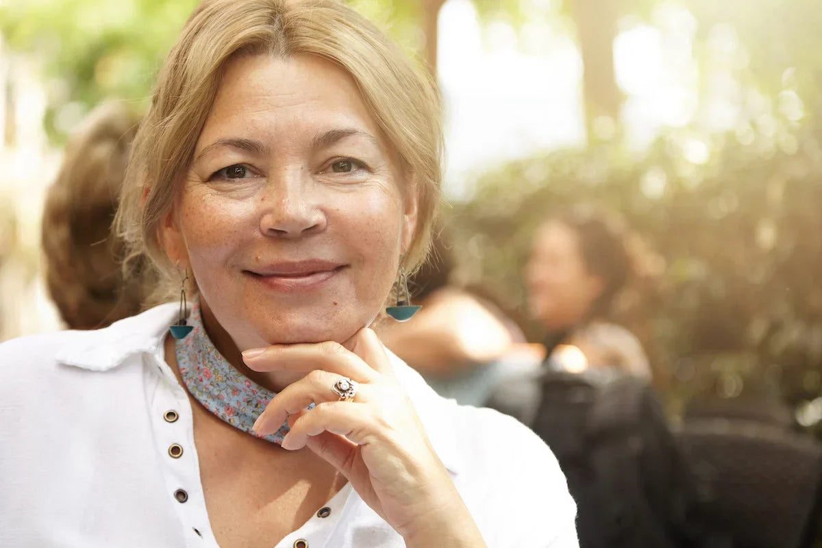 Portrait of a smiling older woman with light hair sitting outdoors, wearing earrings and a floral scarf.