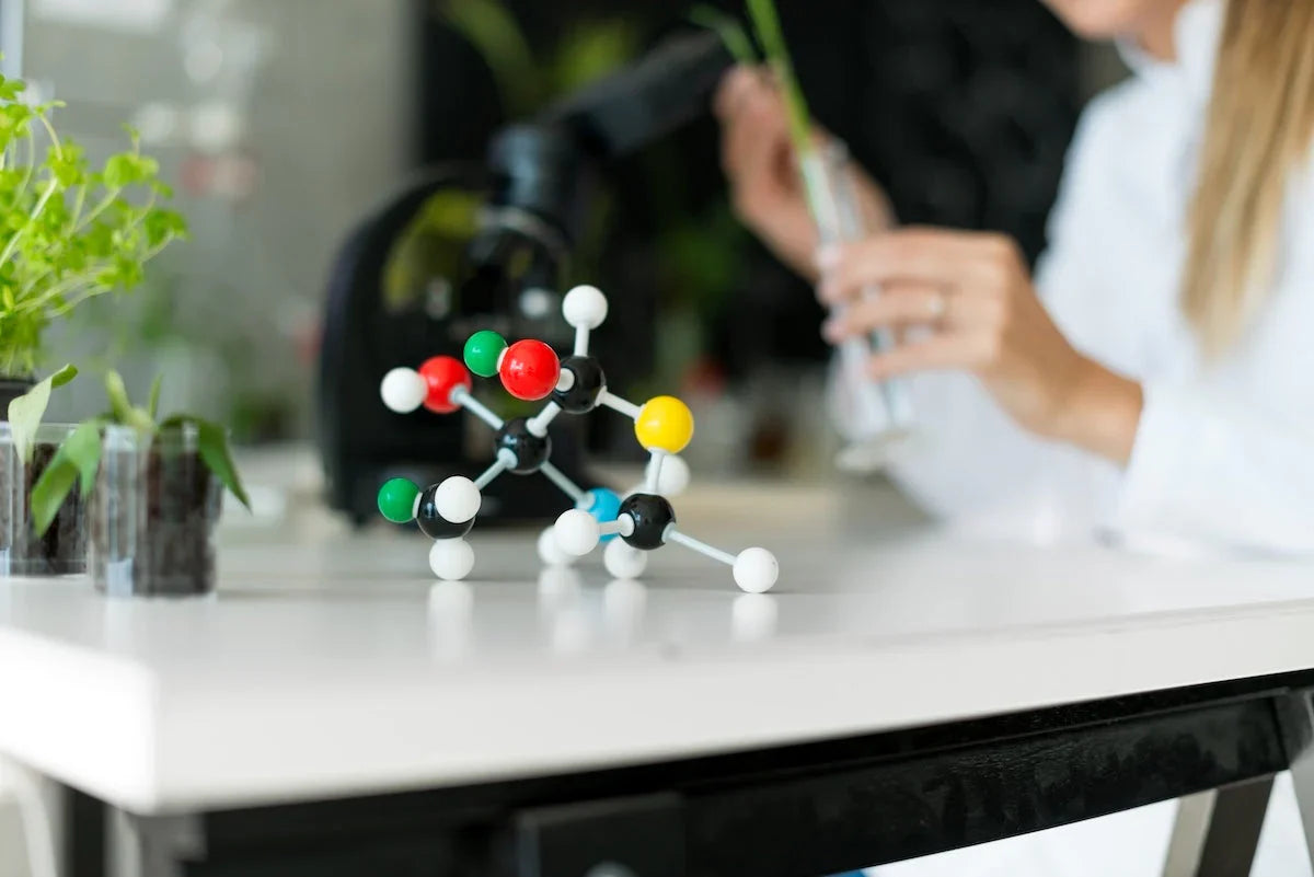 Laboratory scene with a molecular model on the table, plants in containers, and a scientist working with a test tube in the background.