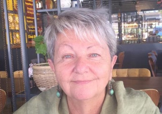 Elderly woman with short gray hair smiling gently at the camera, wearing green earrings and a green shirt, sitting in a cozy cafe setting.