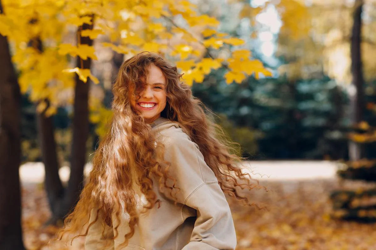 Smiling young woman with long, curly brown hair wearing a beige hoodie, standing outdoors in a park with yellow autumn leaves in the background.
