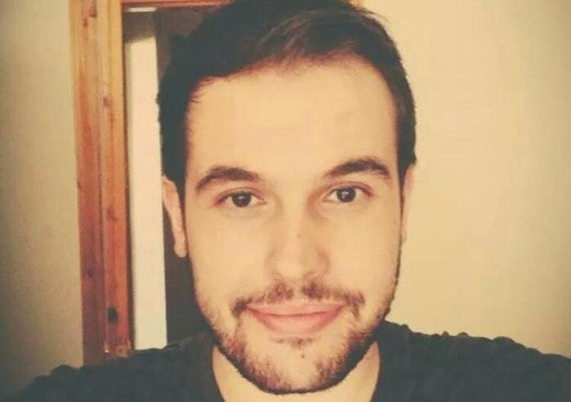 Young man with short dark hair and a trimmed beard smiling slightly at the camera, standing indoors with a wooden door frame visible in the background.