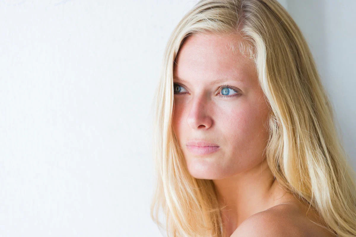 Close-up of a young blonde woman with clear skin and blue eyes, gazing thoughtfully to the side against a light background.